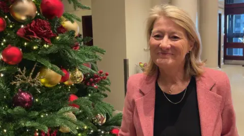 BBC Una mujer con una chaqueta rosa estaba parada junto al árbol de Navidad en el pasillo. ella esta sonriendo
