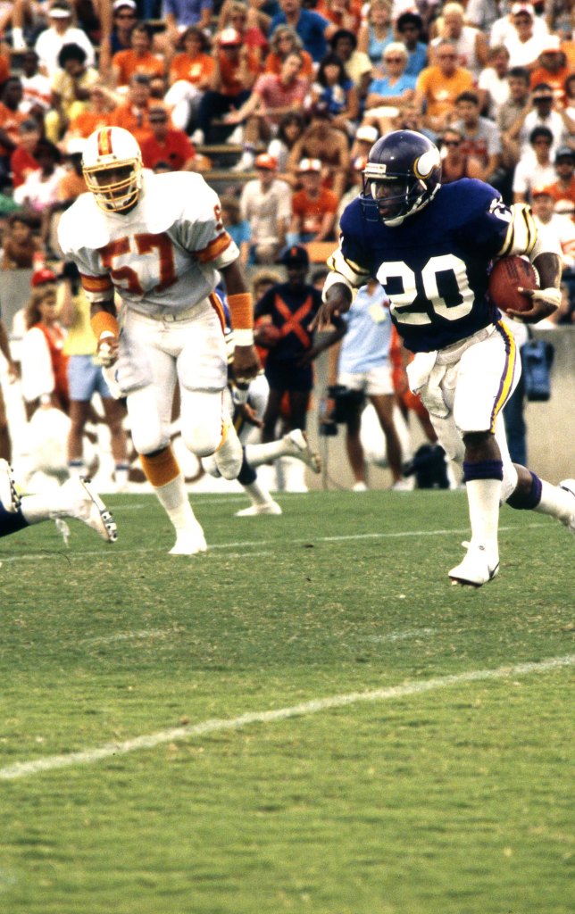 Un jugador de los Minnesota Vikings con una camiseta morada corre con el balón de fútbol número 20, seguido por un jugador de los Tampa Bay Buccaneers con una camiseta blanca con el número 57.