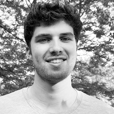 Un joven de pelo corto y rizado y barba clara sonríe al aire libre, vistiendo una camiseta de color claro con árboles al fondo.