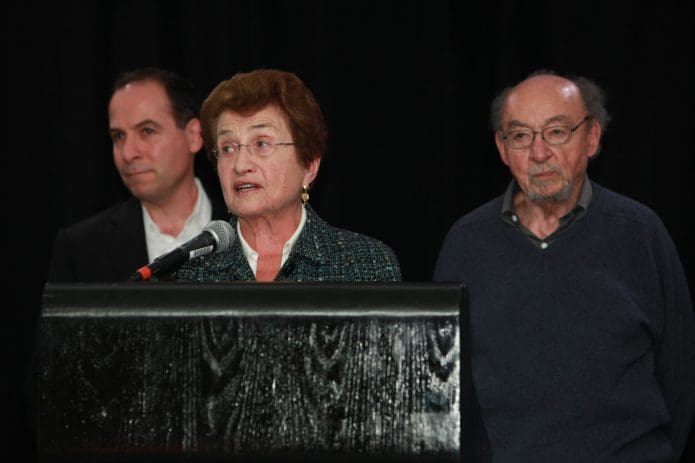 Cora Weiss habla en un servicio conmemorativo del fallecido Saul Landau en 2013. Su hijo Danny (izquierda) y su esposo Peter (derecha) escuchan.