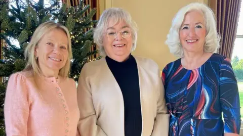 BBC Tres mujeres sonriendo hombro con hombro. Debbie Taylor a la derecha. Tiene el pelo largo y blanco y lleva un vestido azul oscuro.