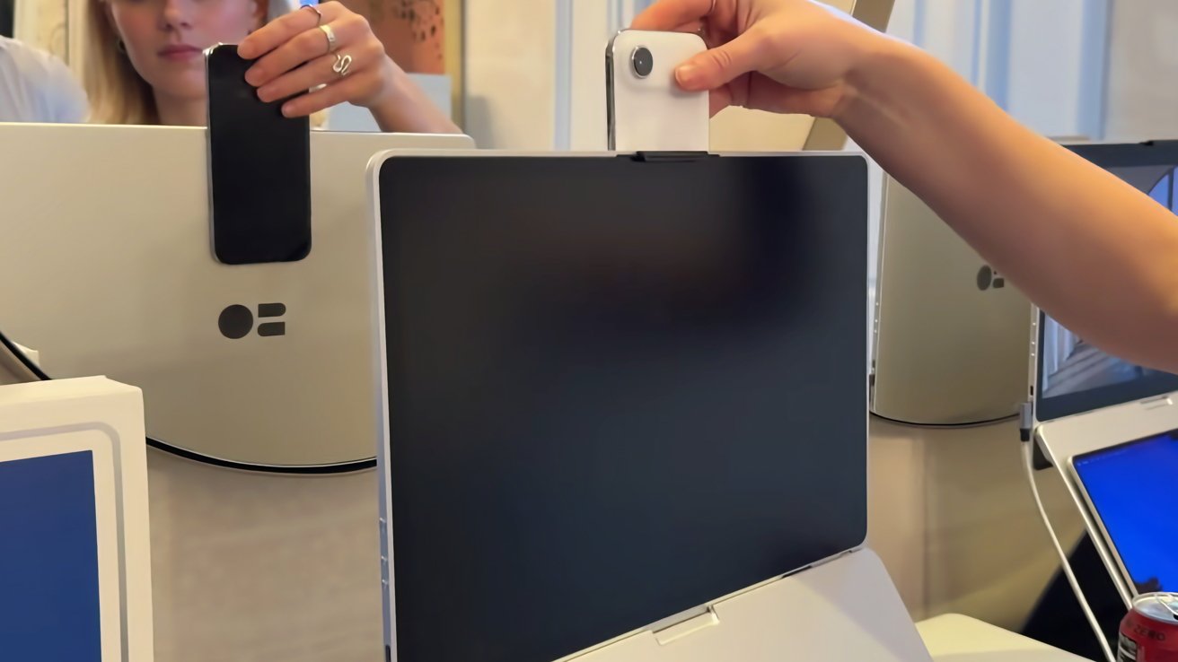 Persona montando un pequeño dispositivo blanco similar a una cámara web encima de un gran monitor de computadora negro, otras pantallas de escritorio y el reflejo de una mujer visible en el fondo