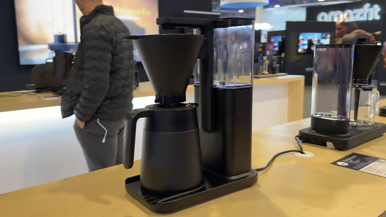 Moderna cafetera de goteo negra con filtro cónico, jarra térmica y depósito de agua clara en un mostrador de madera en una exposición de tecnología, con personas y otros equipos al fondo