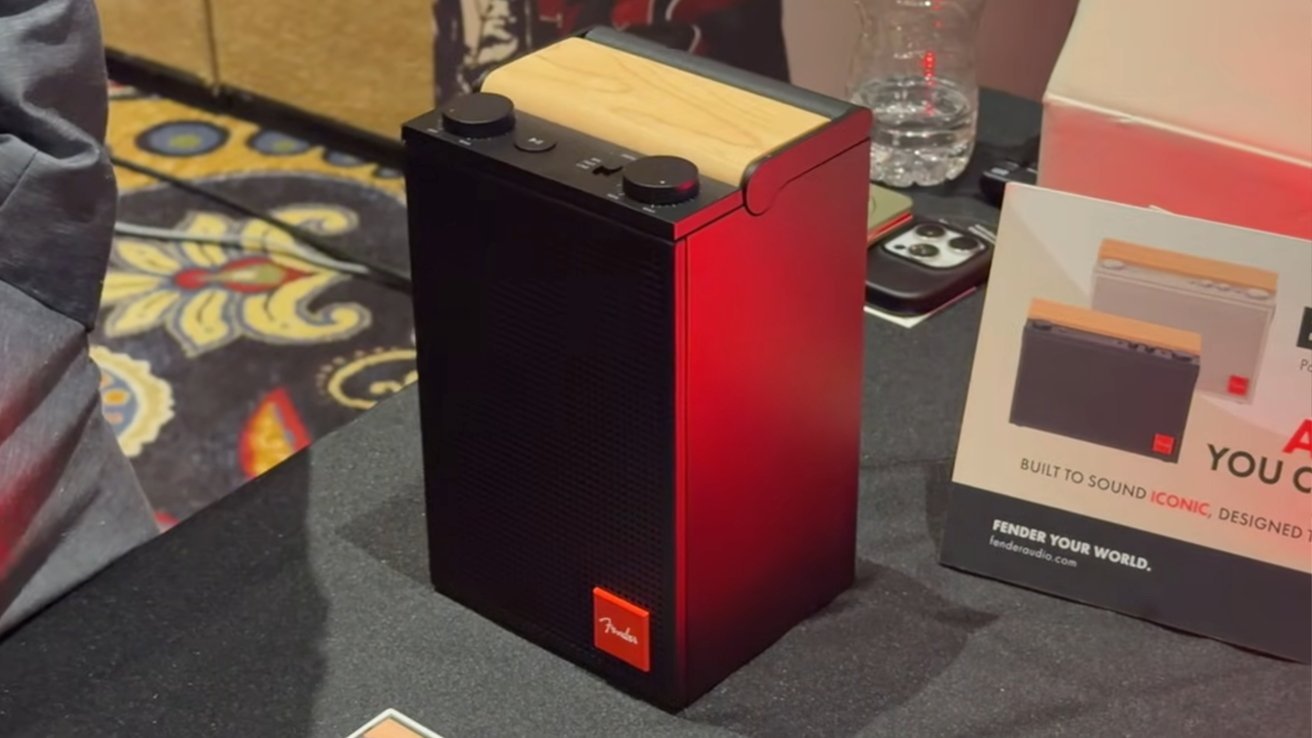 Altavoz inalámbrico Fender alto y rectangular con lados rojos, rejilla negra, panel superior de madera y dos perillas de control negras, exhibido en una mesa junto a materiales promocionales y teléfonos.