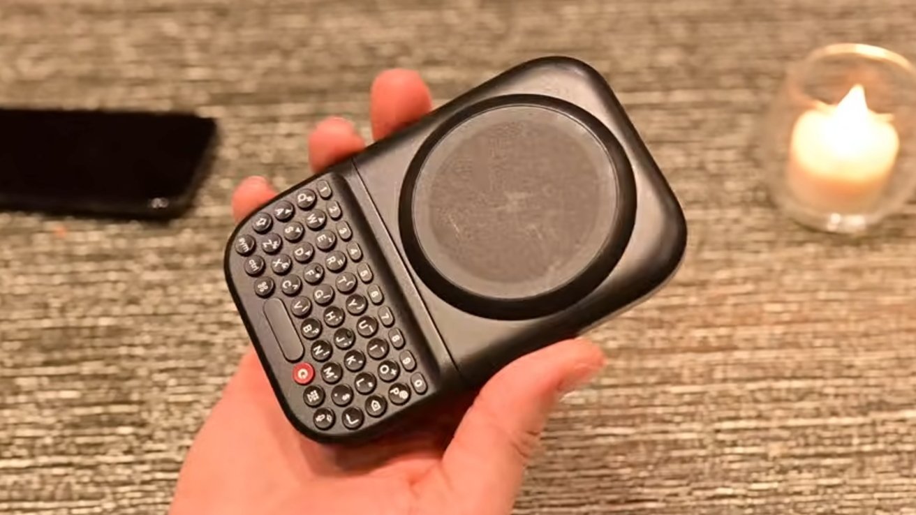 Mano sosteniendo un pequeño dispositivo negro con panel táctil circular y un pequeño teclado QWERTY, un teléfono inteligente apoyado sobre una superficie texturizada y una vela candelita encendida en el fondo
