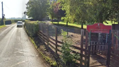 Google Un camino corre a la izquierda de una valla de madera y un seto, con un cartel de escuela detrás de una puerta.