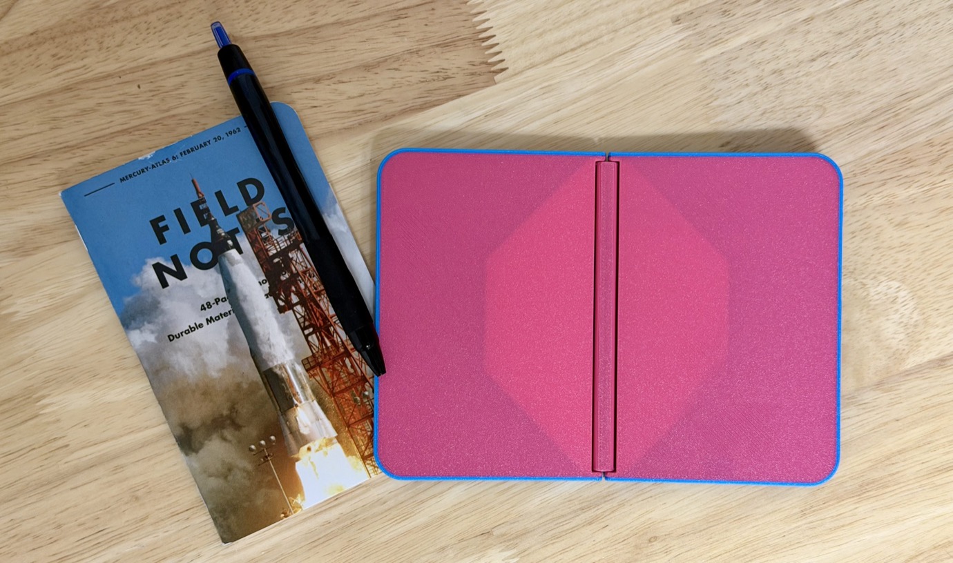 Un cuaderno de notas de campo con tapa rosa y borde azul está abierto sobre una superficie de madera junto a un bolígrafo negro con punta azul y una revista con el lanzamiento de un cohete.