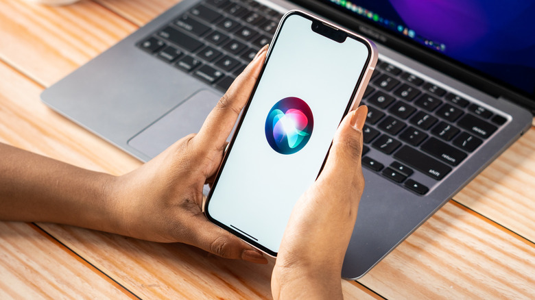 Una persona sostiene un iPhone entre ellos mientras muestra el logotipo de Siri, detrás hay un dispositivo MacBook