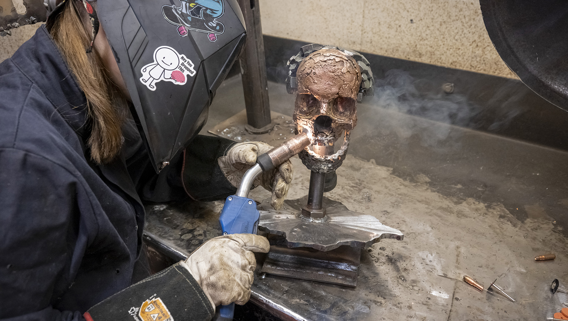 Izzy Chappell usa una máscara de soldadura y guantes de soldadura mientras usa un soplete sobre una escultura de metal con forma de cráneo humano sobre una mesa.