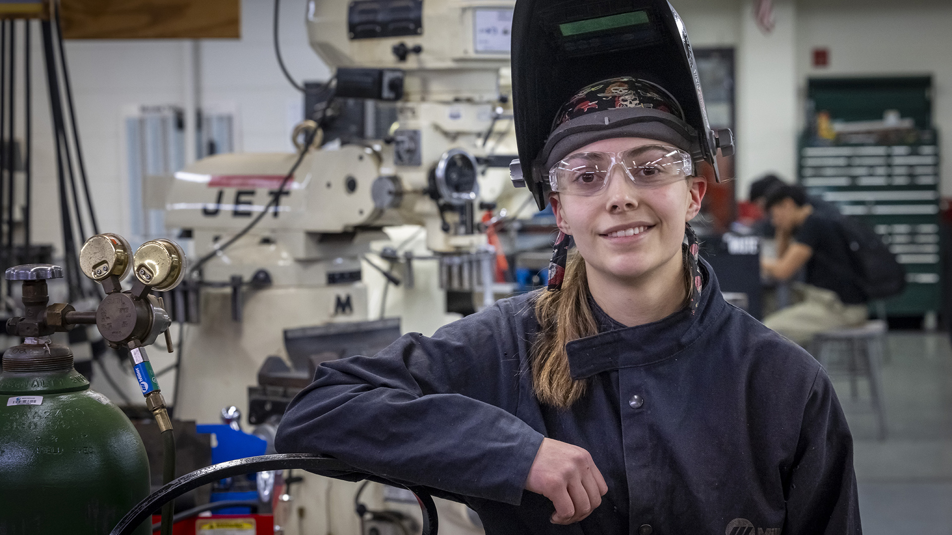 Easy Chappell posa para una fotografía en una sala con equipos de fabricación desenfocados, estantes de herramientas y personas sentadas al fondo mientras usa una máscara de soldadura con el visor levantado y apoyado contra un cilindro de gas de soldadura.